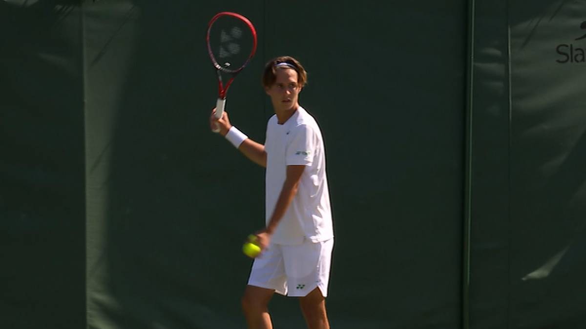 Młody mężczyzna w białej koszulce polo i krótkich spodenkach, trzymający rakietę tenisową w prawej ręce i piłkę tenisową w lewej.