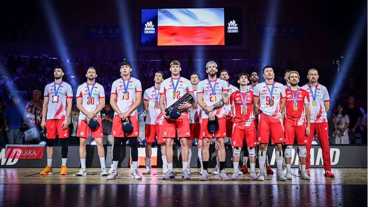 Drużyna siatkarzy Polski w strojach sportowych z medalami na szyjach stoi na scenie, pozując do zdjęcia. W tle wyświetlana jest flaga Polski.