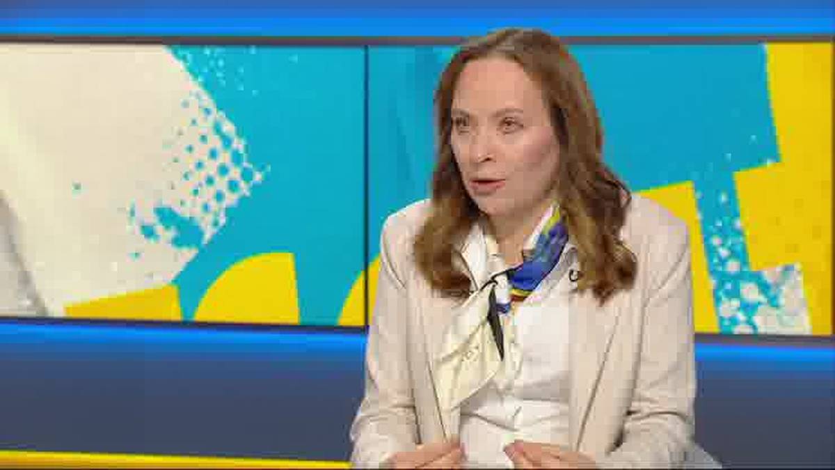 Kobieta w jasnej marynarce i białej koszuli, ubrana w apaszkę z niebieskimi i ż&oacute;łtymi elementami, siedzi i m&oacute;wi. Tło stanowią dwa ekrany z abstrakcyjnymi, geometrycznymi wzorami w kolorach niebieskim i ż&oacute;łtym.