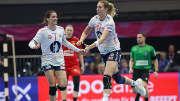 Dwie zawodniczki piłki ręcznej w białych strojach cieszą się po udanej akcji, podając sobie ręce.