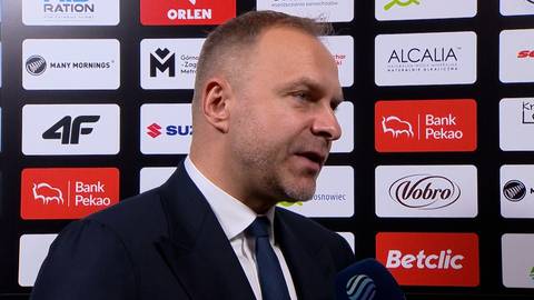 Mężczyzna w garniturze z krawatem m&oacute;wi do mikrofonu, na tle widoczne są logotypy sponsor&oacute;w.