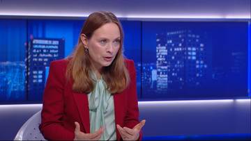 Kobieta w czerwonej marynarce i jasnej bluzce z kokardą, m&oacute;wiąca z rękami uniesionymi w geście rozmowy.