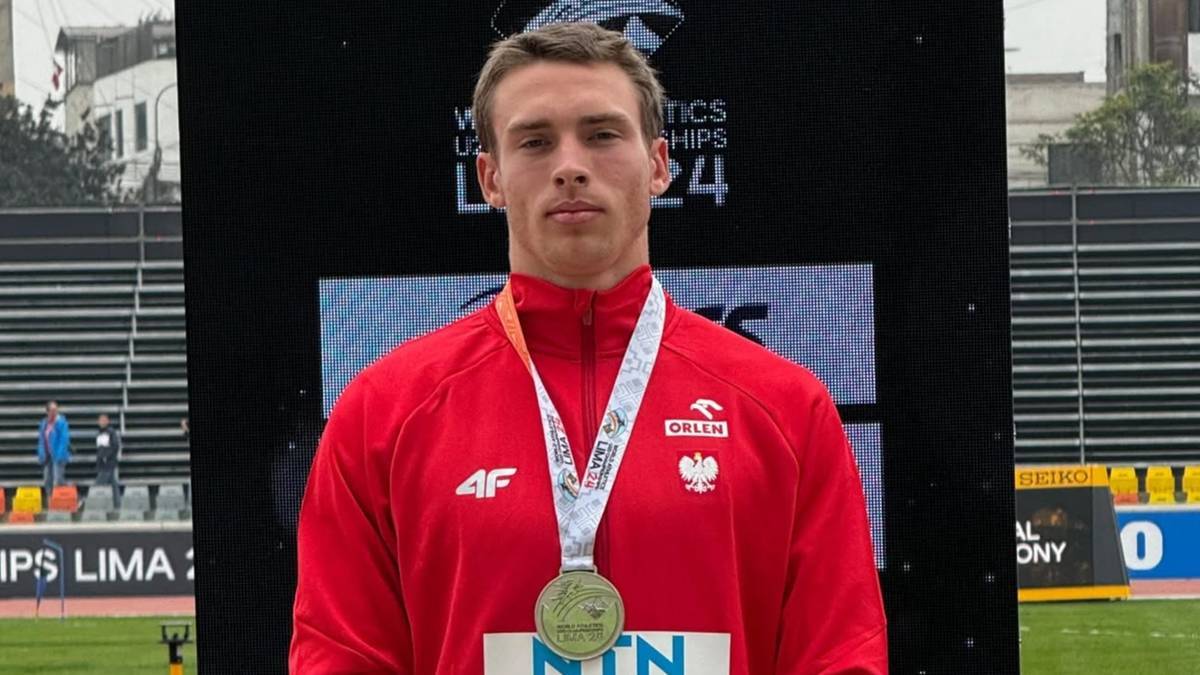 Młody mężczyzna w czerwonej bluzie sportowej z orłem na piersi, noszący złoty medal na szyi, pozuje na tle bandy reklamowej z napisem "WORLD ATHLETICS CHAMPIONSHIPS LIMA 24".