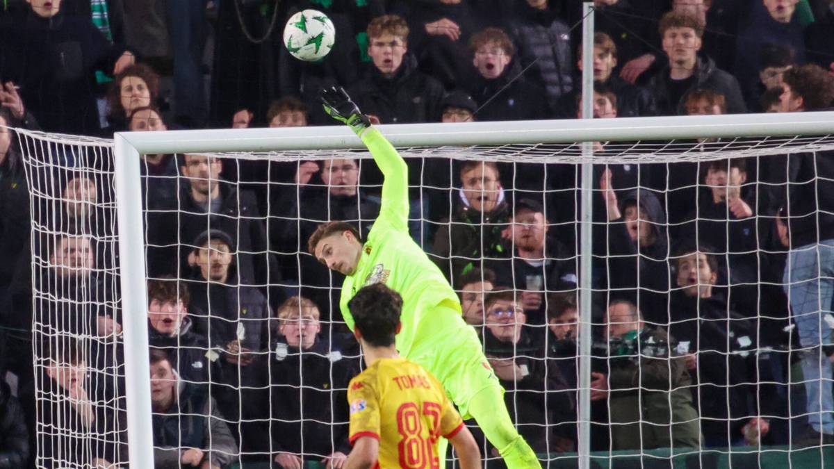 Bramkarz w zielonym stroju skacze do obrony strzału piłki, a napastnik w ż&oacute;łto-czerwonym stroju obserwuje sytuację.