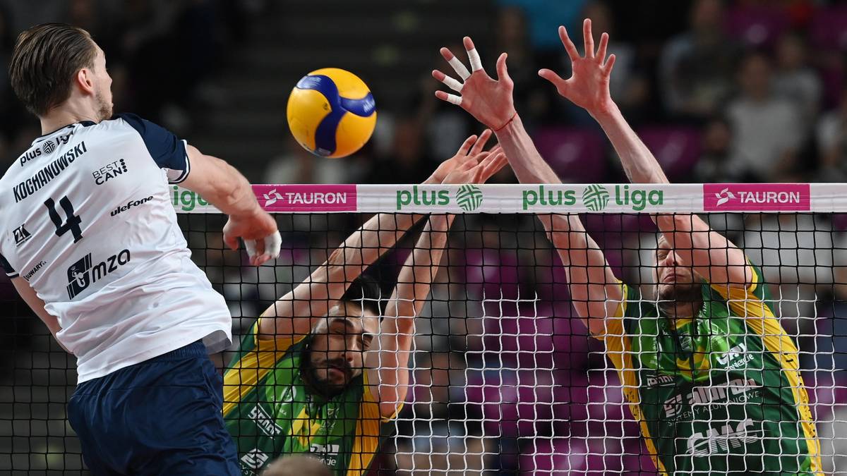 Siatkarze w akcji, jeden z nich w białej koszulce próbuje zaatakować piłkę, podczas gdy dwaj gracze w zielonych koszulkach starają się ją zablokować nad siatką.