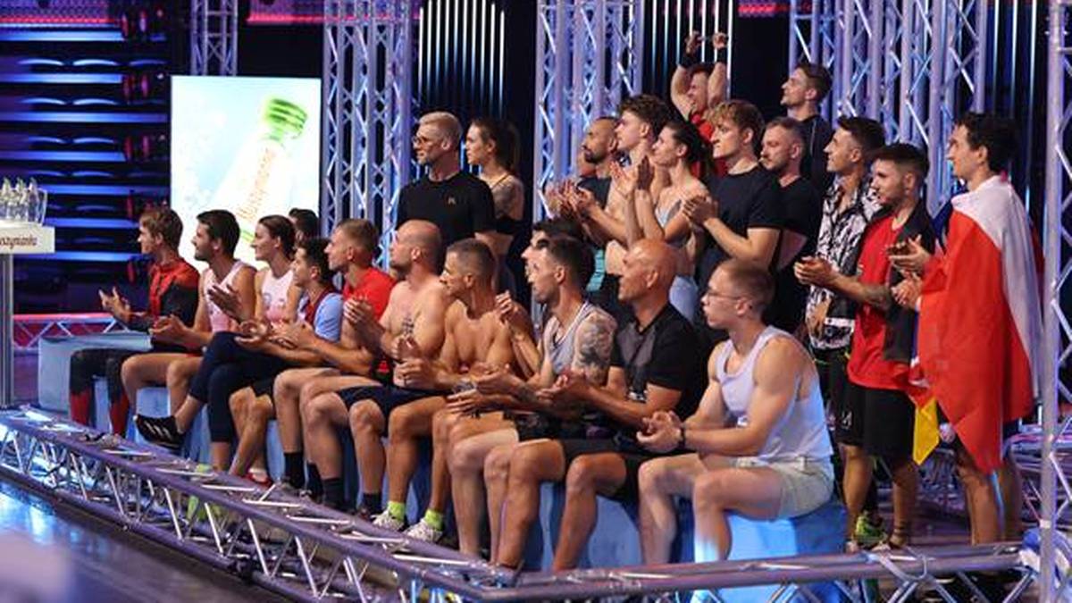 Grupa młodych ludzi siedzi na ławce przed areną sportową. Niektórzy machają, inni patrzą w stronę kamery. Wszyscy są ubrani w sportowe stroje.