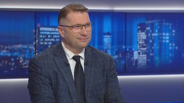 Przemysław Czarnek w garniturze i krawacie siedzi w studiu telewizyjnym, uśmiecha się. W tle widoczne są nocne panoramy miast na ekranach.