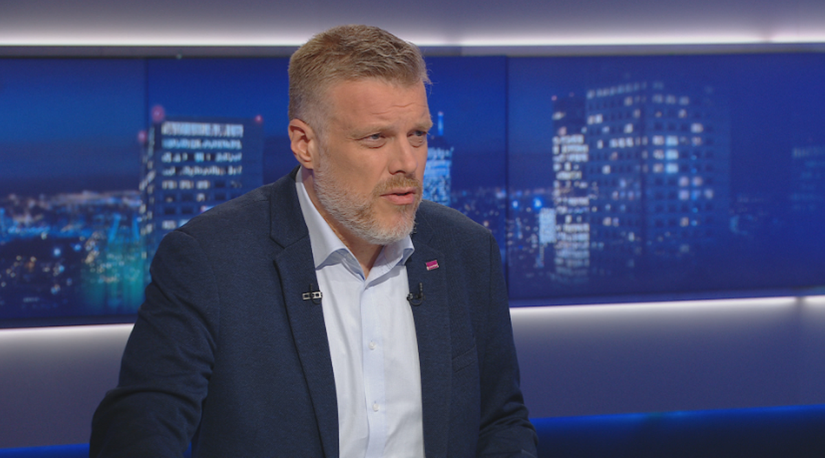 Mężczyzna w średnim wieku z brodą, ubrany w granatową marynarkę, wypowiada się w studiu telewizyjnym na tle z widokiem na nocne miasto.
