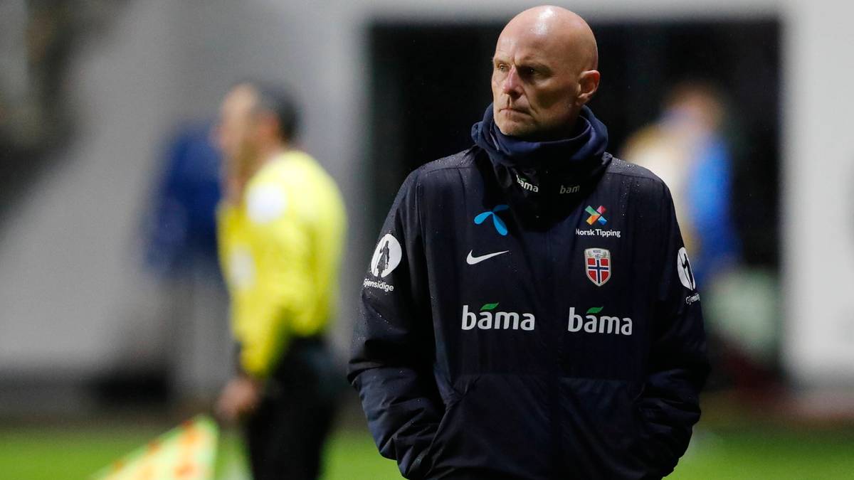 Mężczyzna w średnim wieku, łysy, z poważnym wyrazem twarzy, ubrany w ciemną kurtkę sportową z widocznym logo Norwegii, stoi na boisku piłkarskim, w tle niewyraźne postacie.