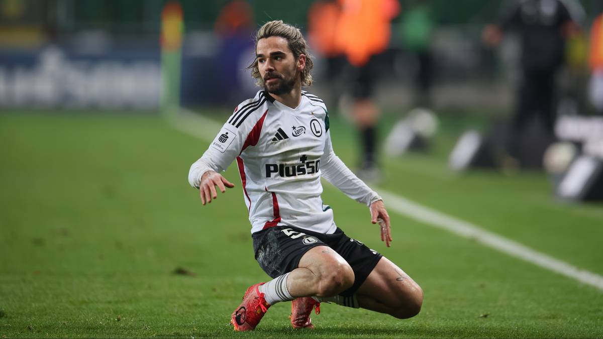 Piłkarz Legii Warszawa w białej koszulce i czarnych spodenkach klęczy na boisku.