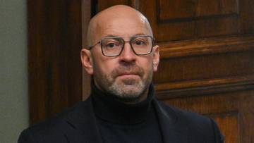 Mężczyzna w garniturze stoi przed dwuskrzydłowymi drewnianymi drzwiami, zapinając marynarkę.