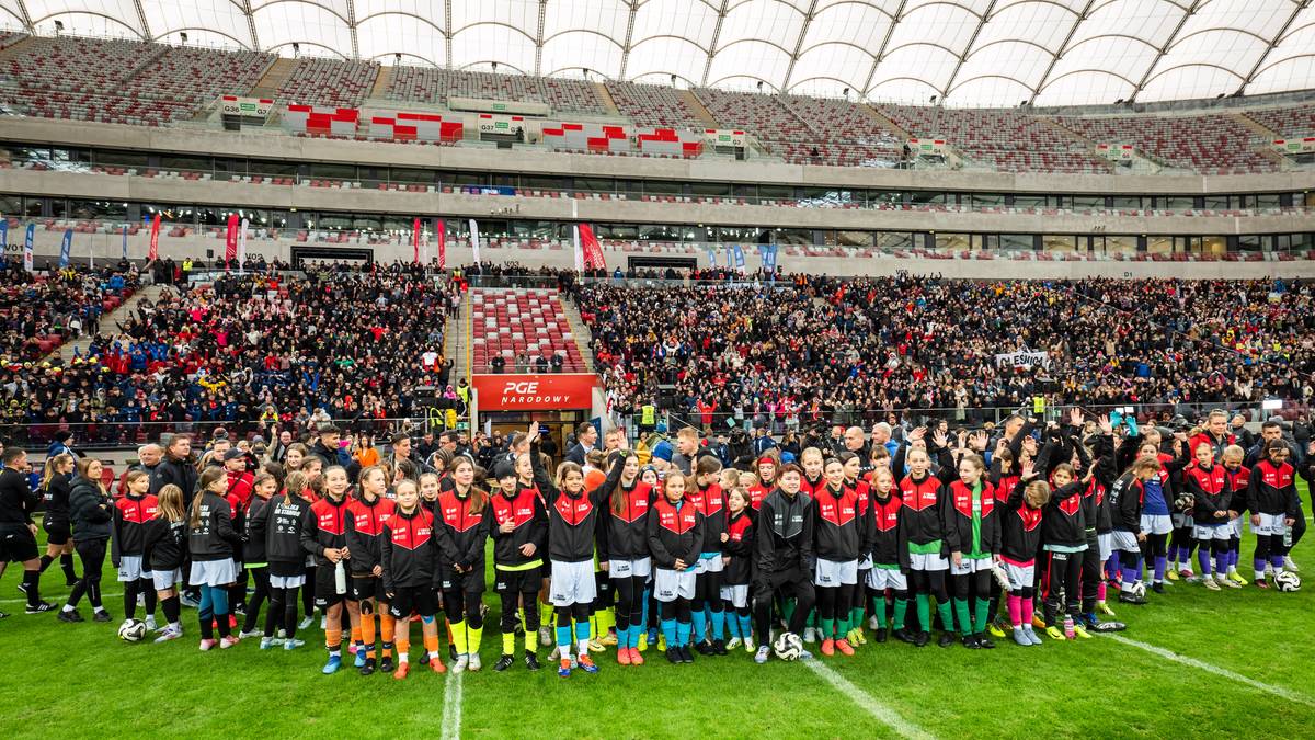 Grupa dziewcząt w strojach piłkarskich stoi na zielonej trawie przed trybunami stadionu wypełnionymi publicznością.