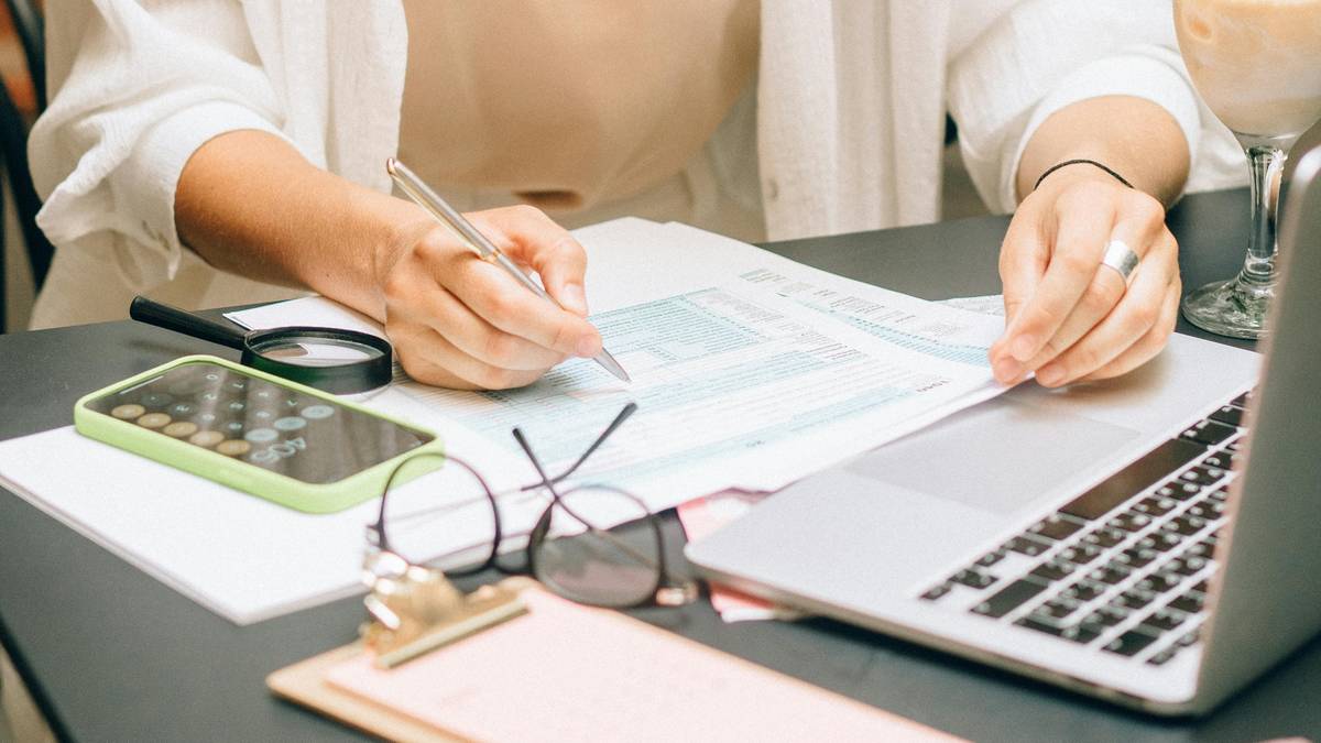 Ręce osoby piszącej na dokumentach, obok laptop, kalkulator i lupa.