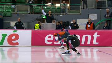 Zawodnicy łyżwiarstwa szybkiego przygotowują się do startu, jeden z sędziów trzyma pistolet startowy w górze.