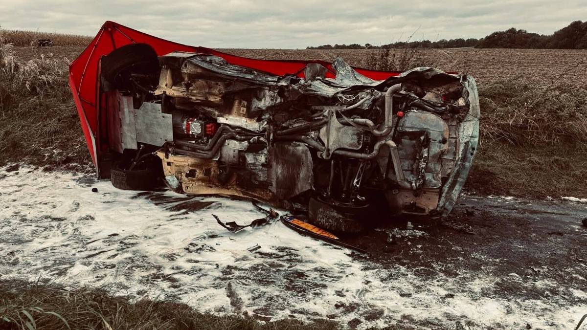 Spalony samochód rajdowy leżący na boku na poboczu drogi, otoczony zniszczeniem.