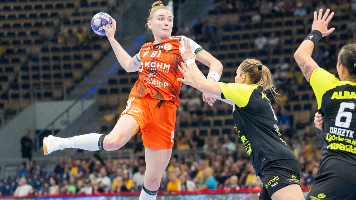 Zawodniczka KGHM Zagłębia Lubin w pomarańczowym stroju, z numerem 91, wykonuje rzut do bramki. Broniąca ją zawodniczka drużyny przeciwnej pr&oacute;buje ją zablokować.