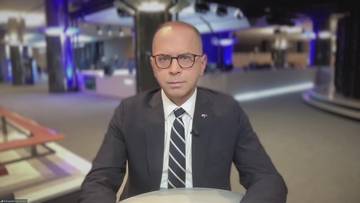 Mężczyzna w okularach i garniturze siedzi przed rozmytym tłem studia telewizyjnego.