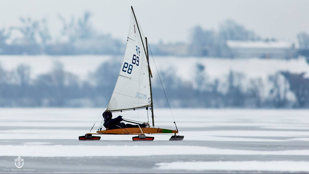 Bojer z numerem 28 na żaglu porusza się po zamarzniętym jeziorze, widziany z profilu. Zawodnik siedzi wewnątrz łodzi.