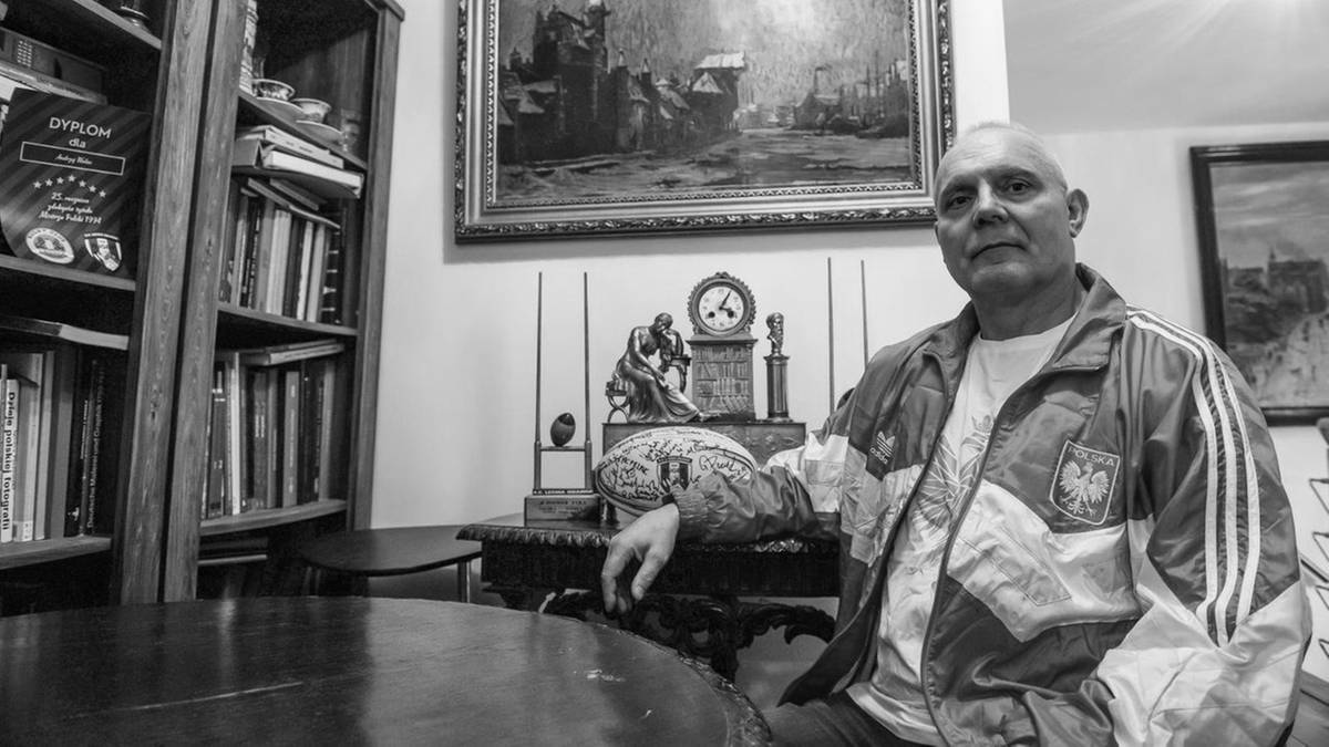 Portret mężczyzny w ok. 50. roku życia, siedzącego na tle wnętrza z książkami i obrazami.