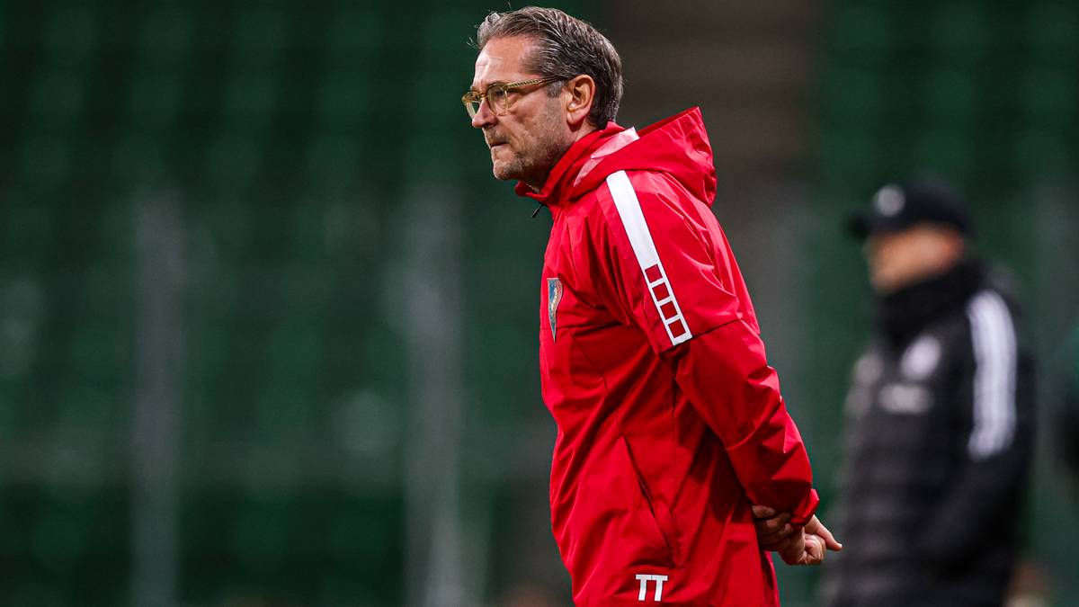 Mężczyzna w czerwonej kurtce treningowej z białymi pasami na ramionach, w okularach, stoi na boisku piłkarskim i obserwuje grę.