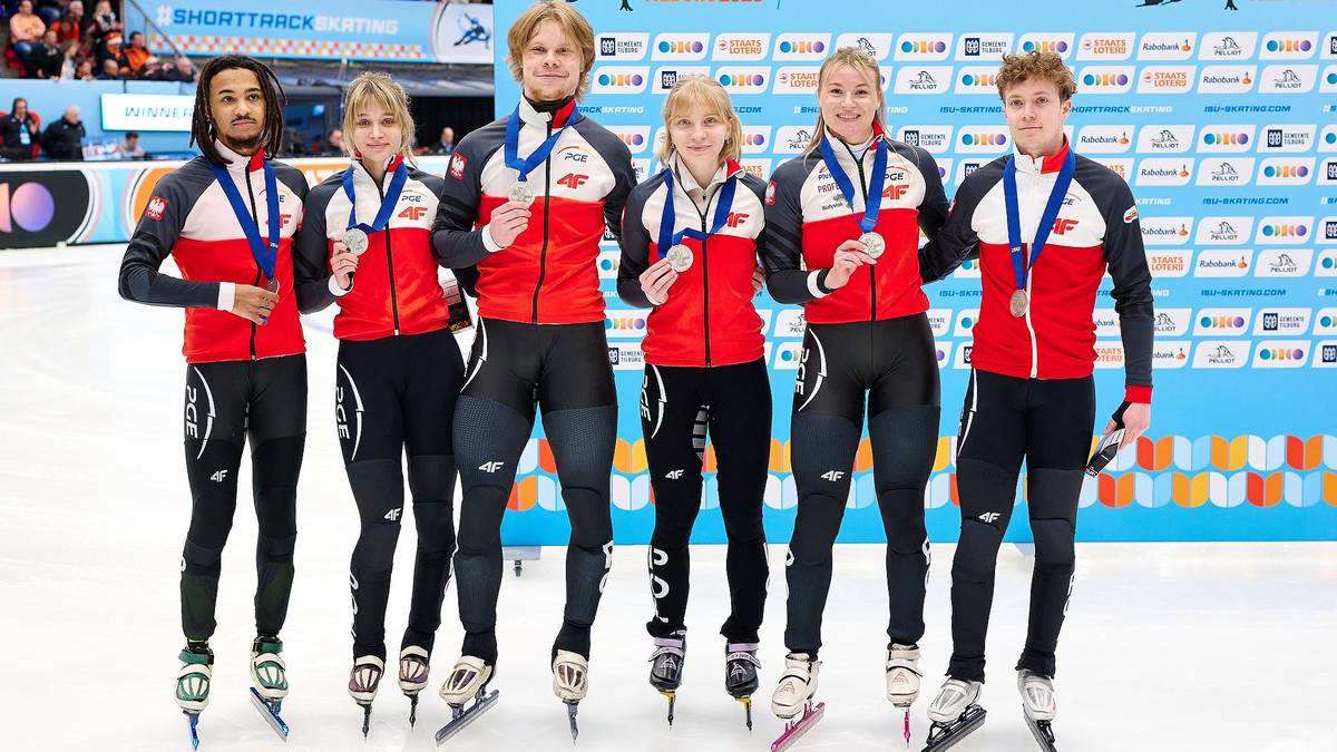 Drużyna łyżwiarzy szybkich w strojach sportowych z medalami na szyjach pozuje do zdjęcia na lodowisku.