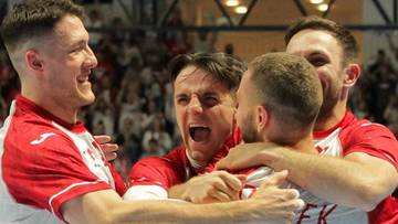 Pięciu mężczyzn w białych strojach sportowych z czerwonymi detalami, obejmuje się i świętuje radośnie zwycięstwo.