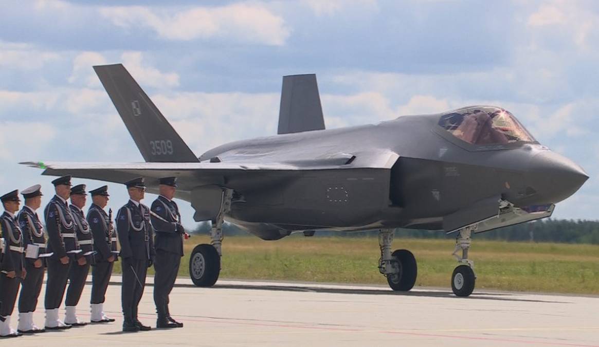Myśliwiec F-35 "Husarz" z widocznym numerem bocznym 3005, stojący na pasie startowym, obok niego w szeregu stoi grupa polskich wojskowych w galowych mundurach.
