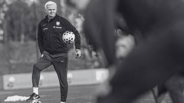 Trener piłkarski w stroju sportowym z piłką przy nodze na boisku.