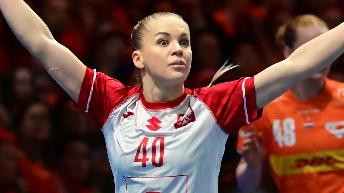 Piłkarka ręczna w białej koszulce z numerem 40 i czerwonymi spodenkami świętuje zdobytego gola z uniesionymi rękami, na trybunach widać kibiców.