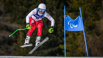 Narciarz w kombinezonie z białym kaskiem i goglami zjeżdża po stoku narciarskim, trzymając kije.