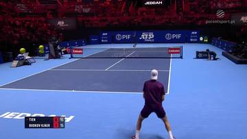 Tenisista Learner Tien przygotowuje się do serwisu na korcie tenisowym podczas meczu z Nicolai Budkov Kjaer. Widoczni są kibice na trybunach i logo ATP Tour.