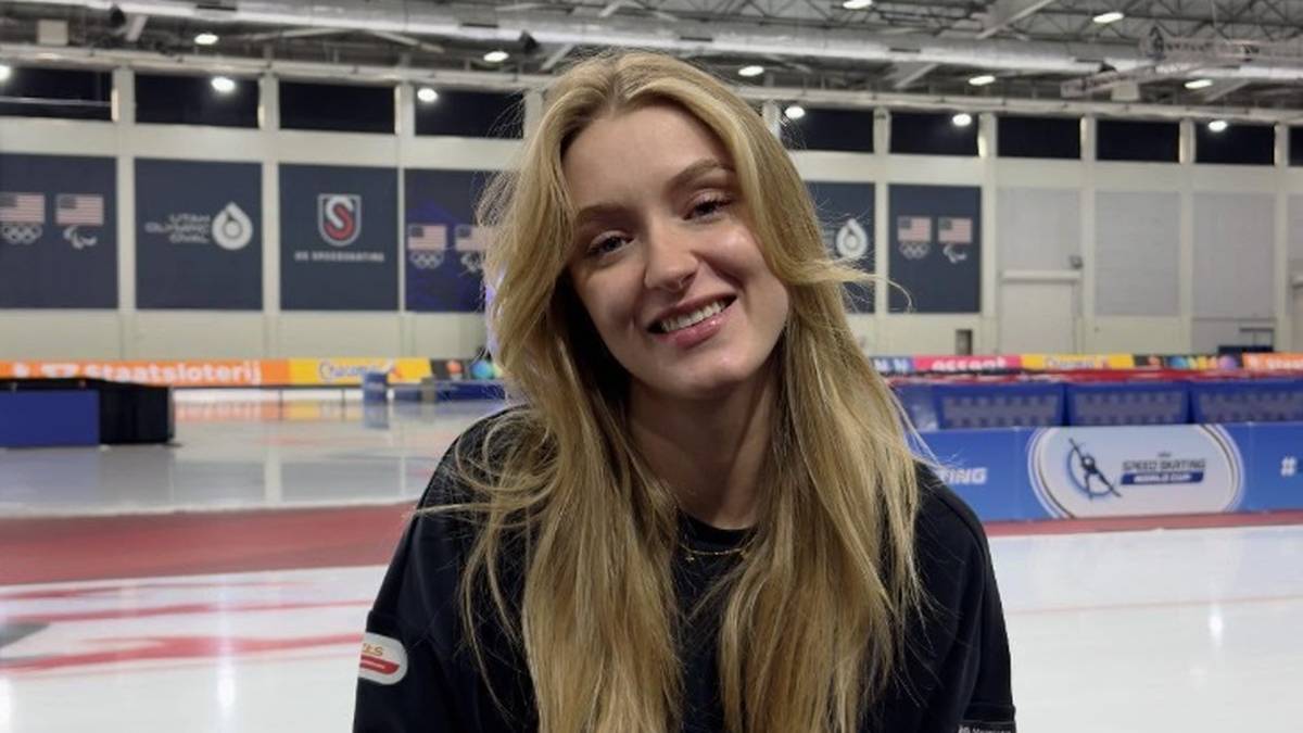 Młoda kobieta o blond włosach siedzi uśmiechnięta na lodowisku, w tle widoczne są elementy infrastruktury sportowej.