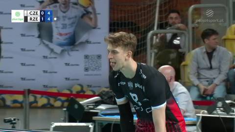 Młody siatkarz w czarnej koszulce z czerwonymi akcentami i numerem 8 na plecach, koncentruje się na grze podczas meczu siatk&oacute;wki. W tle widoczny wynik spotkania na tablicy.