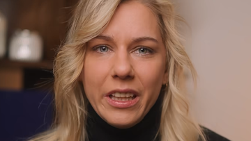 Zbliżenie na twarz blondwłosej kobiety z niebieskimi oczami, m&oacute;wiącej z otwartymi ustami. Kobieta ma na sobie czarny golf.