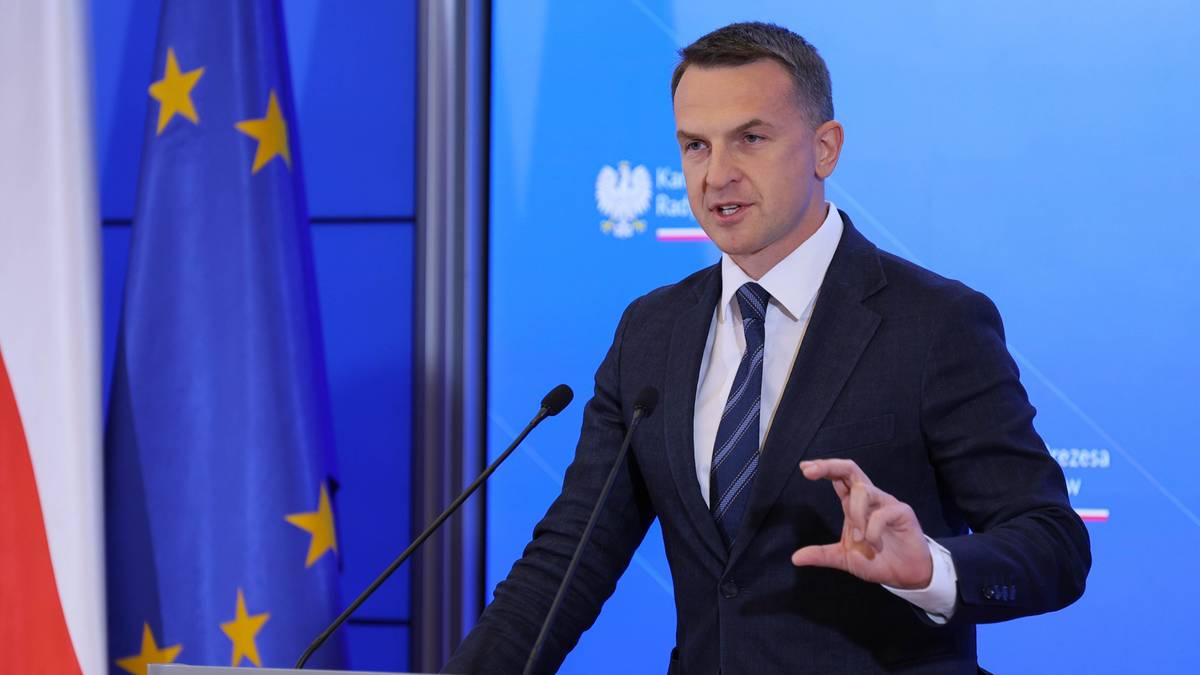 Mężczyzna w garniturze przemawia przy m&oacute;wnicy, obok flagi Polski i Unii Europejskiej.