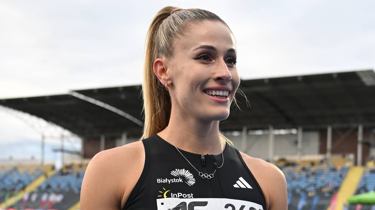 Portret młodej lekkoatletki z blond kucykiem, uśmiechniętej, w stroju sportowym z logo "Białystok" i "InPost", na tle trybun stadionu.