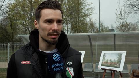 Mężczyzna w czarnej kurtce z kapturem, trzymający mikrofon, stoi na tle otwartego terenu sportowego z ławkami i stojącym na statywie obrazem przedstawiającym park.