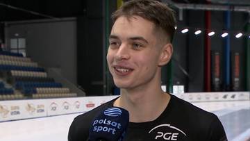 Młody mężczyzna w czarnej koszulce z logo PGE i Polsat Sport udziela wywiadu na lodowisku.