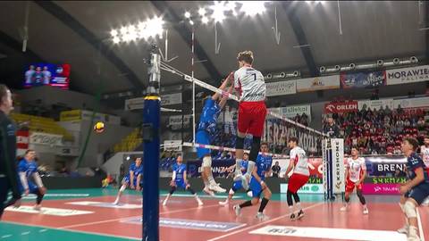 Siatkarz w czerwonym stroju wyskakuje do ataku nad siatką, podczas gdy zawodnik w niebieskim stroju próbuje go zablokować.