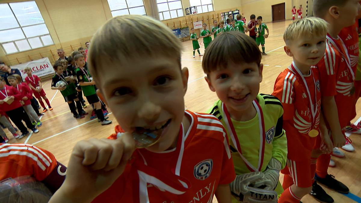 Dwóch chłopców w strojach sportowych z medalami na szyjach, jeden z nich żuje medal, uśmiechają się do kamery, w tle hala sportowa z uczestnikami turnieju.