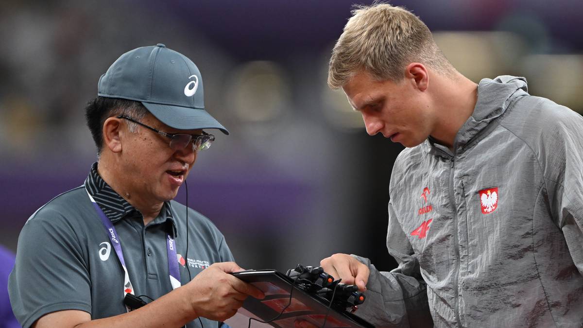 Trener w czapce i okularach z tablicą w ręku rozmawia z blondwłosym sportowcem w szarej bluzie z orzełkiem na piersi.