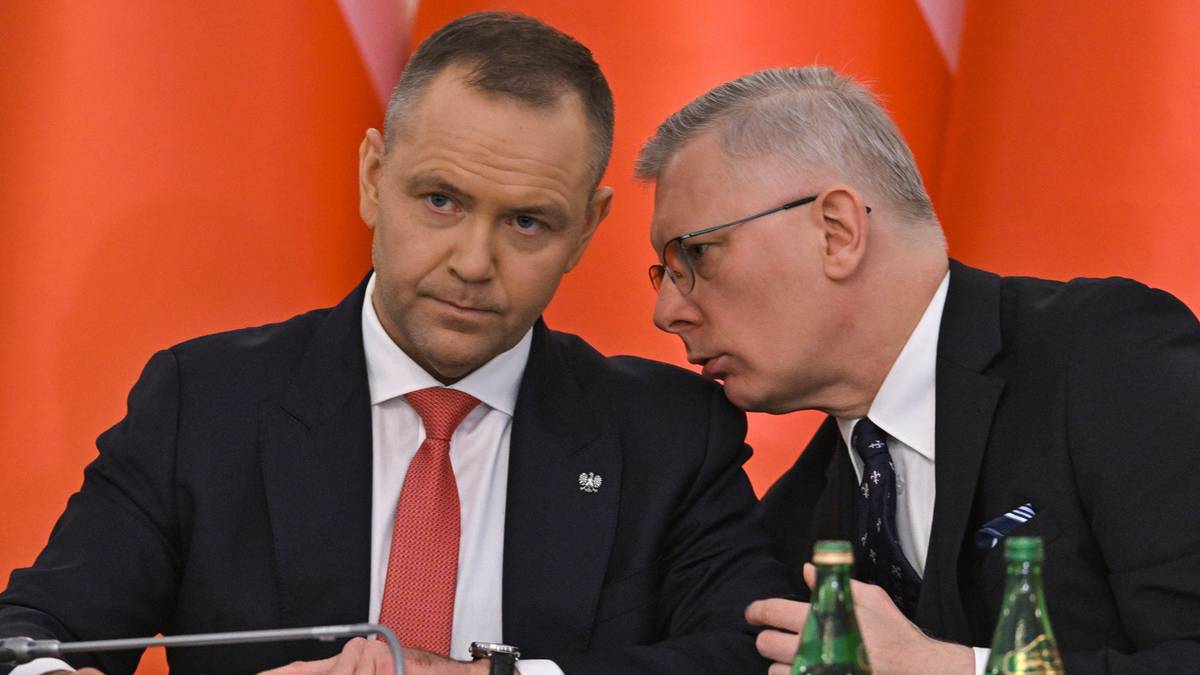 Dw&oacute;ch mężczyzn w formalnych strojach siedzi przy stole, jeden z nich pochyla się i m&oacute;wi do drugiego.