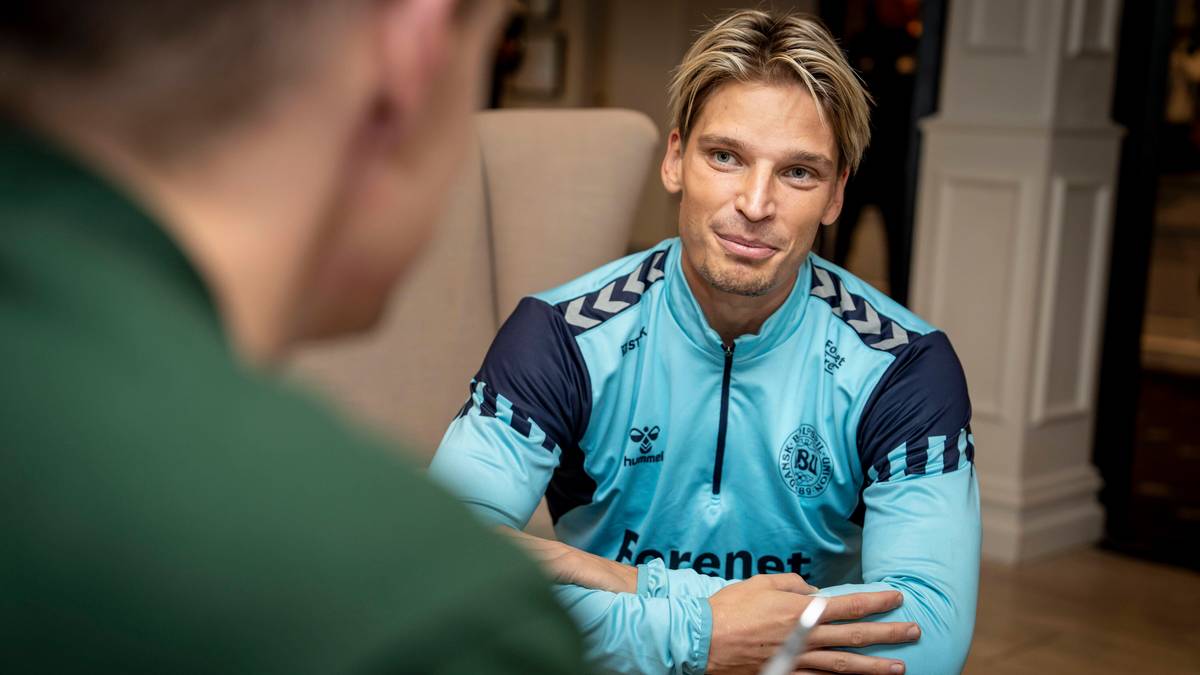 Mężczyzna w niebieskiej koszulce piłkarskiej siedzi przy stole i rozmawia z kimś, kogo twarz jest niewidoczna. Ma blond włosy i patrzy prosto przed siebie.