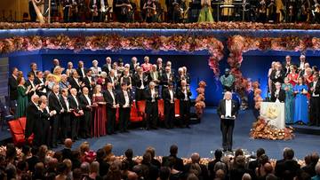 Scena z uroczystości wręczenia Nagród Nobla, na której szwedzki monarcha wręcza dyplomy laureatom w obecności publiczności.