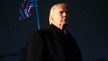 Donald Trump w ciemnym płaszczu z amerykańską flagą w tle.