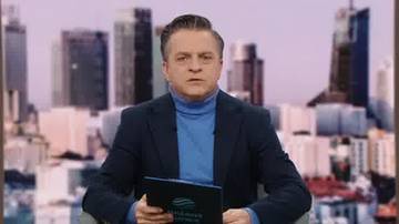 Mężczyzna w ciemnym garniturze i niebieskim golfie siedzi przed ekranem wyświetlającym panoramę miasta z wieżowcami.