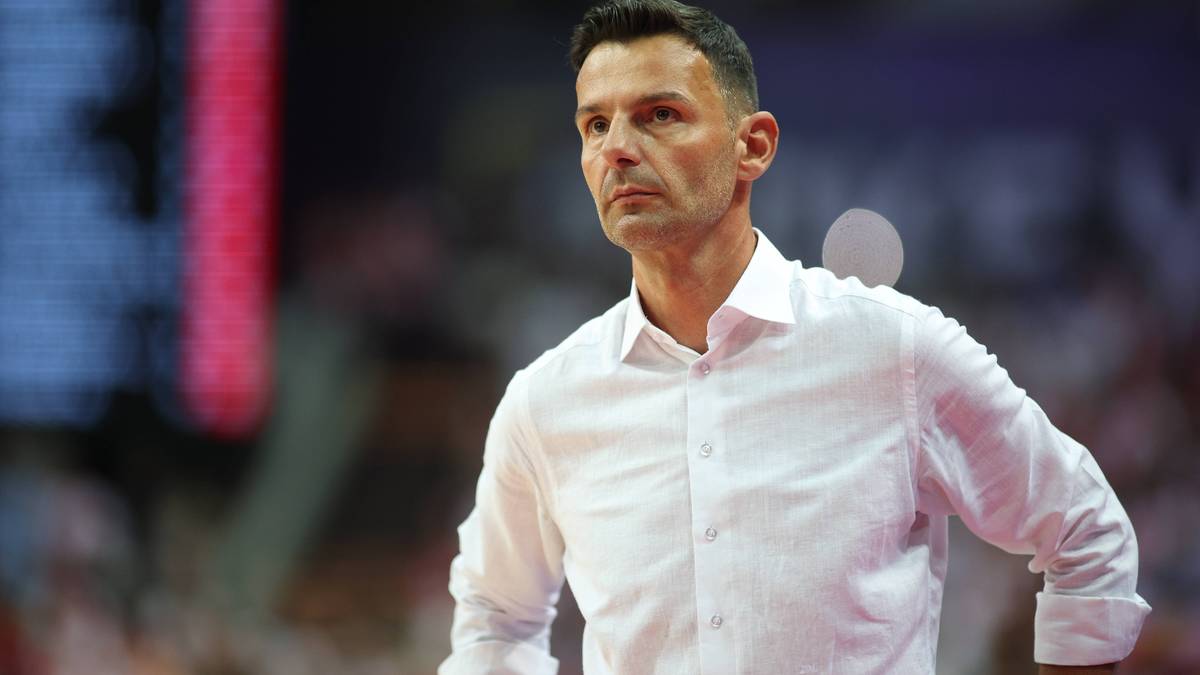 Mężczyzna w białej koszuli, prawdopodobnie trener sportowy, spogląda w bok z poważną miną.