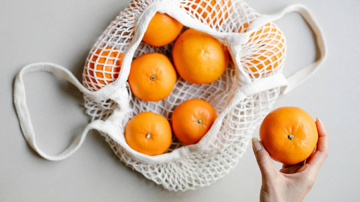 Kilka pomarańczowych mandarynek w białej siatkowej torbie, obok ręka trzymająca jedną mandarynkę.
