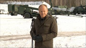 Premier Donald Tusk przemawia przed mikrofonem, w tle widoczne wozy bojowe i zaśnieżony teren.