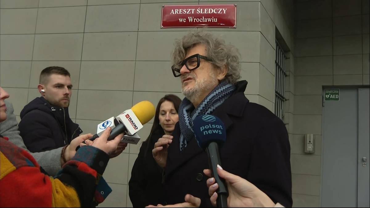 Janusz Palikot z okularami i szalikiem, otoczony przez dziennikarzy z mikrofonami, na tle budynku z napisem "Areszt Śledczy we Wrocławiu".